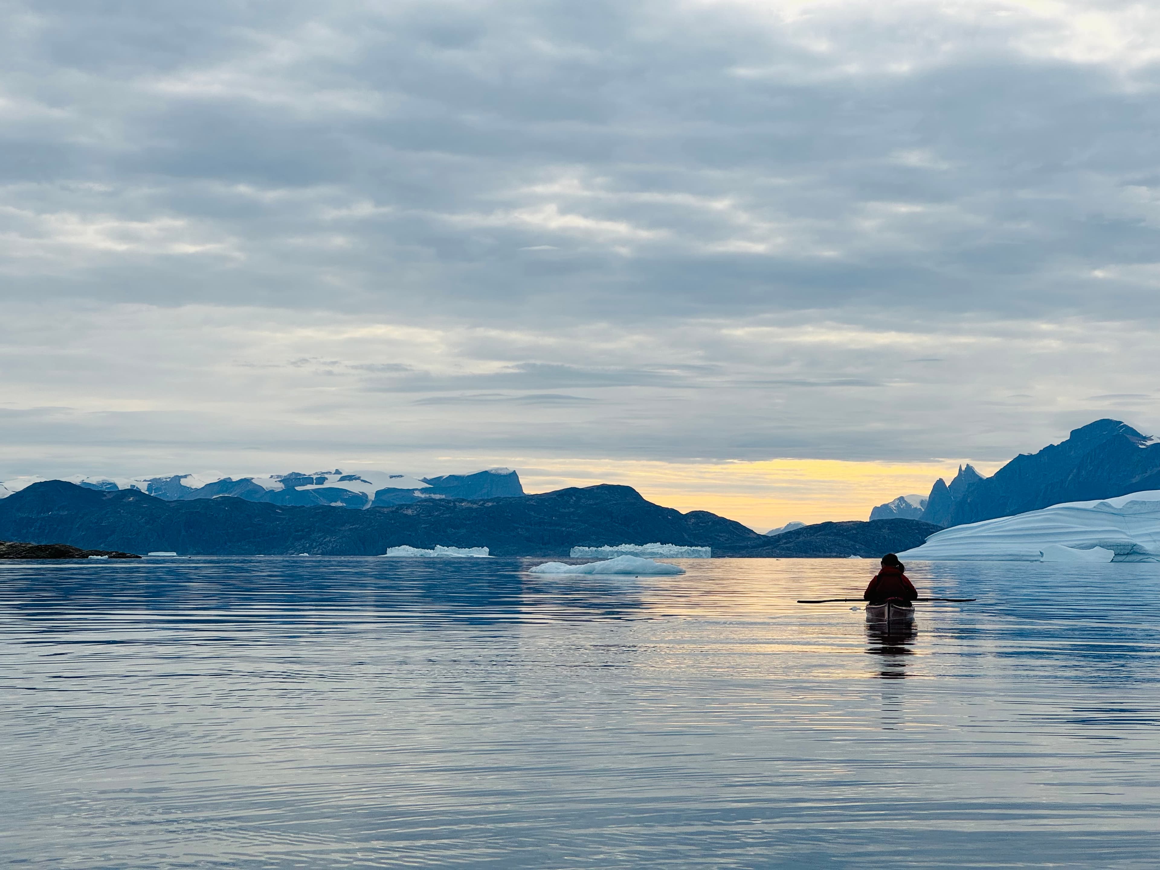 A kayaker on a calm fjord at sunset, icebergs and mountains on the horizon.